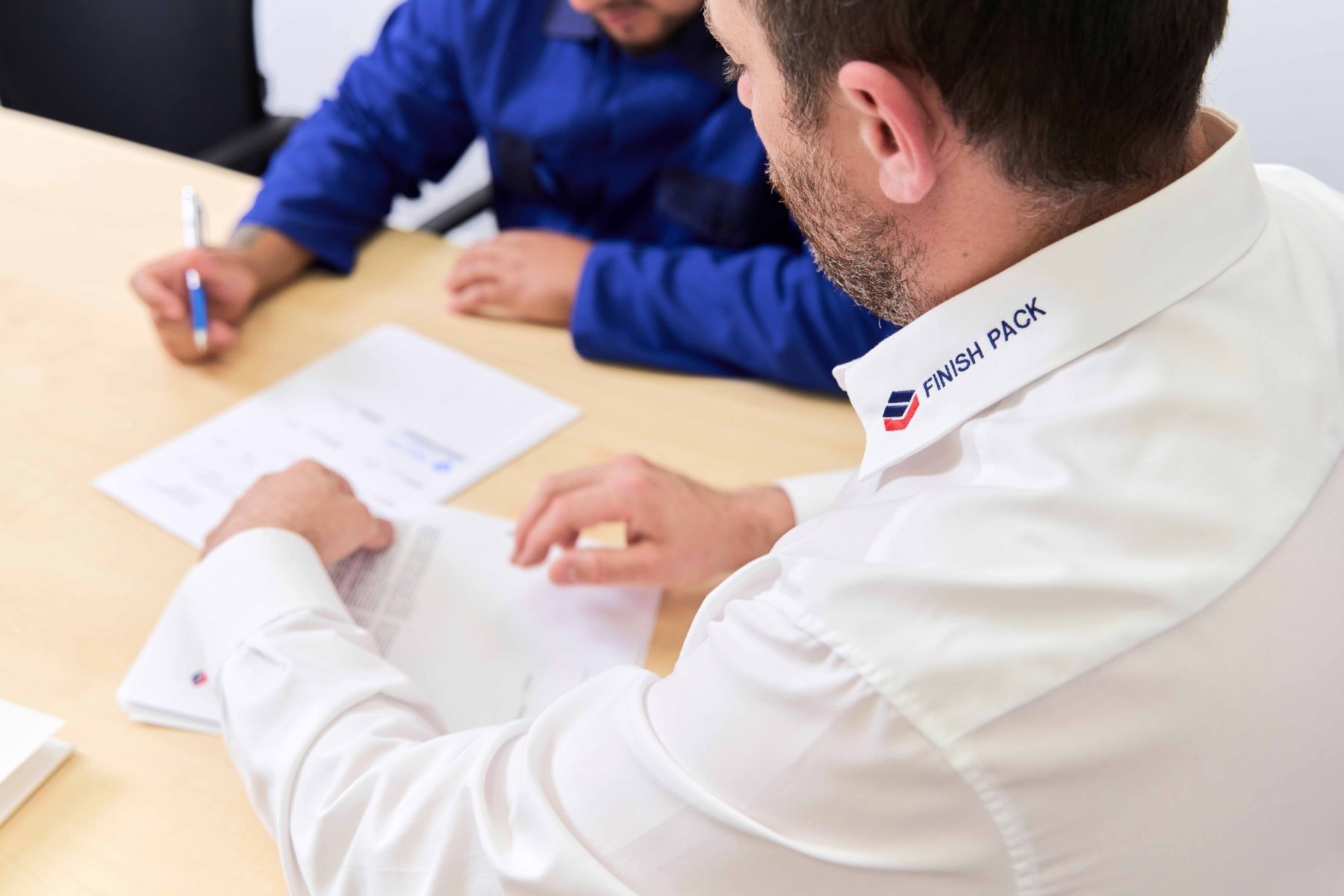 Zwei Männer sitzen an einem Tisch und bearbeiten Papiere, einer trägt ein weißes Hemd mit einem Logo am Kragen.