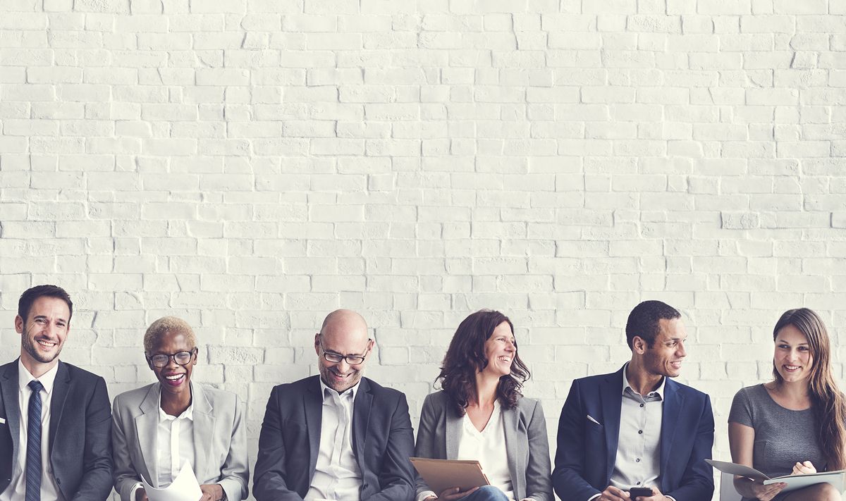 Sechs Bewerber*innen sitzen nebeneinander vor einer weißen Ziegelwand und lachen dabei.